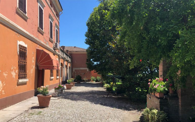 In questa foto si può vedere il retro della casa vista dal cancello d'ingresso, si nota il verde rigoglioso e nell'ombra un dondolo a due posti con il tettuccio fatto di tessuto, comodo per dondolarsi e sentire il vento accarezzarti la faccia.